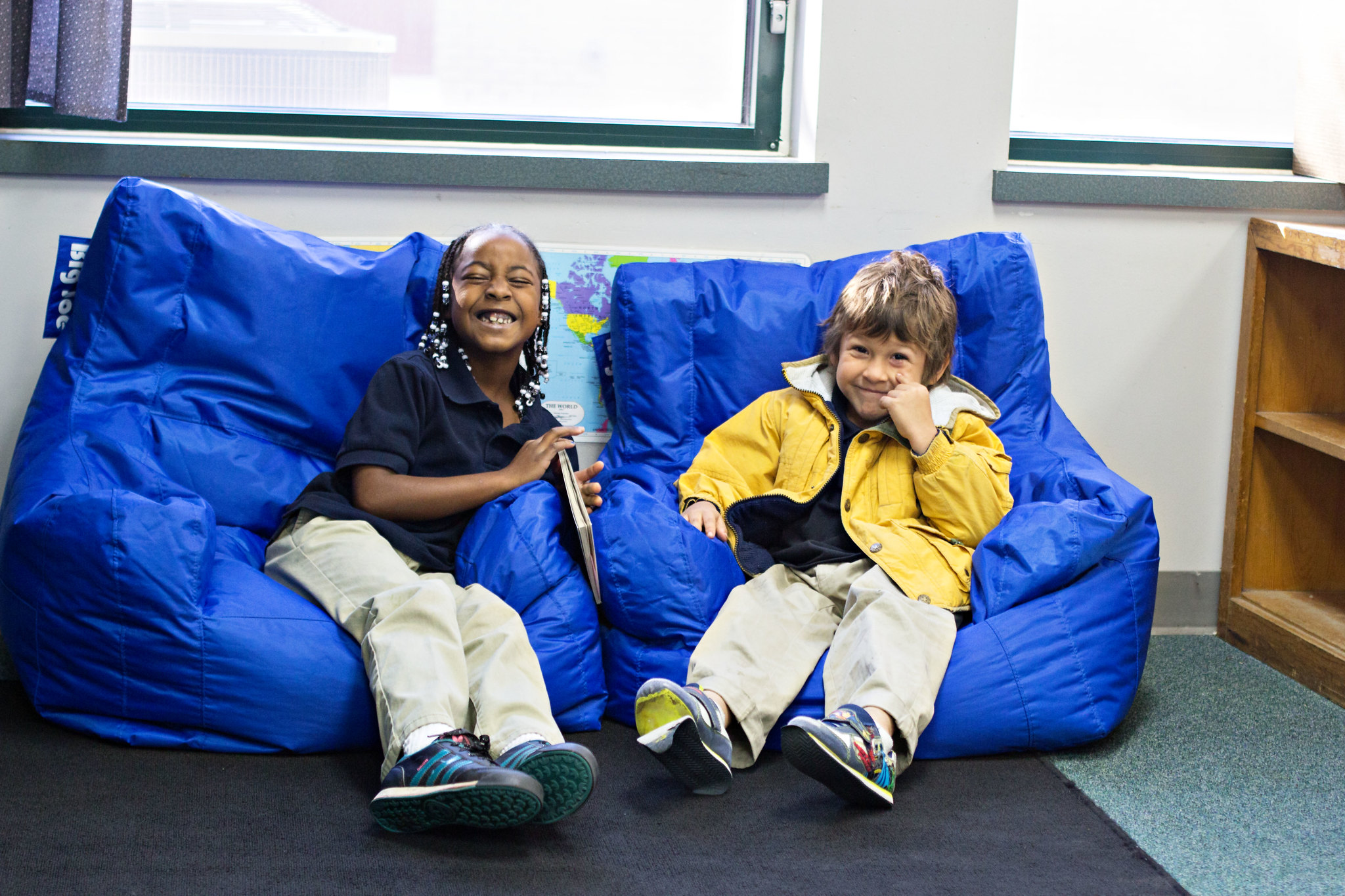 two students, one Black and one White, sit on blue bean bag chairs and laugh together. one student holds a book