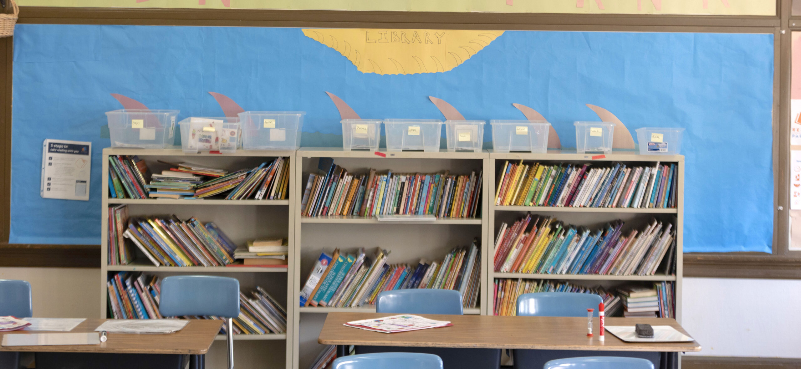 bookshelves against a wall holding colorful picture books, three empty desks sit in front of them