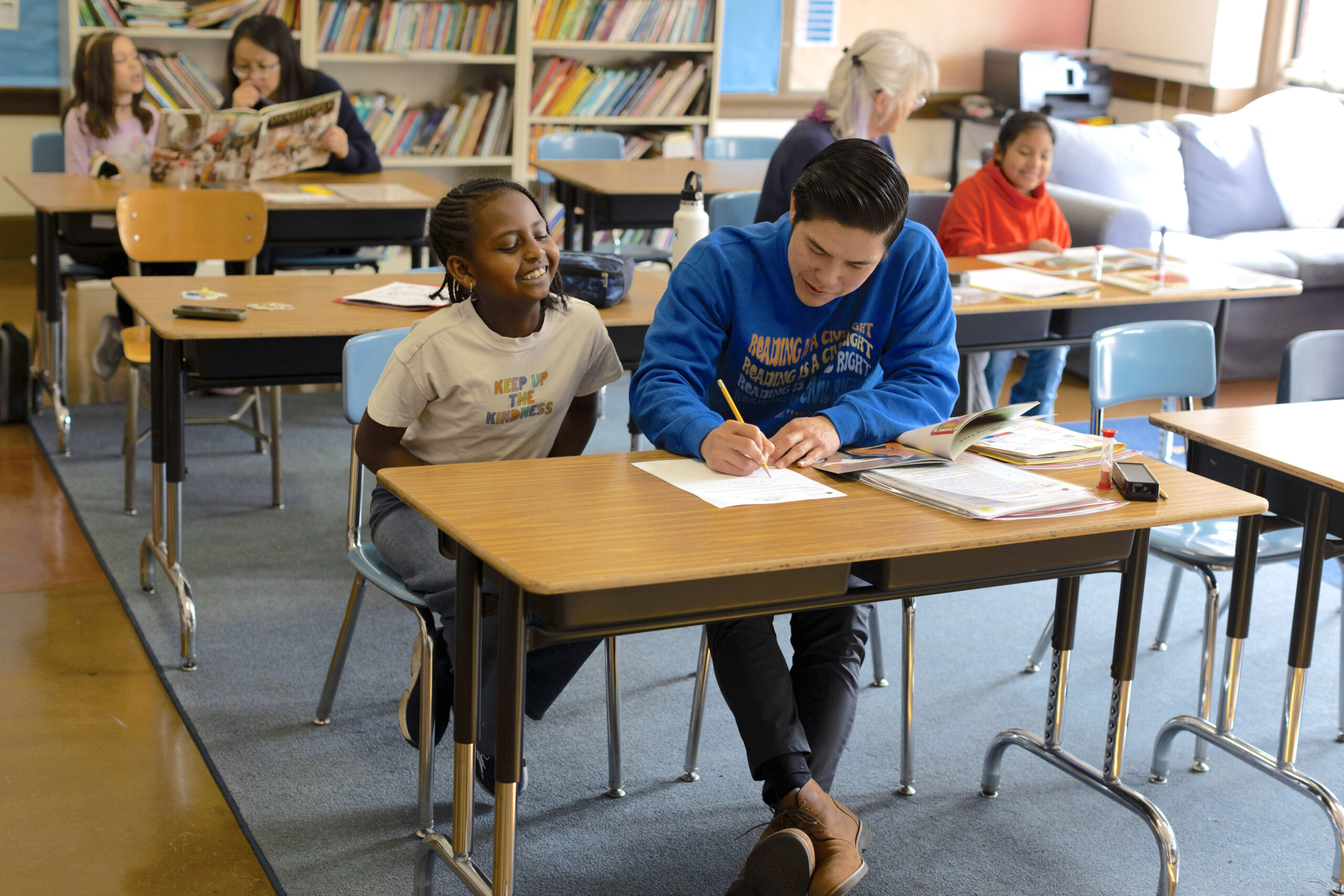 a Reading Partners reading center filled with tutor-student pairs, the tutor-student pair in the forefront are smiling and the tutor is writing something on a piece of paper