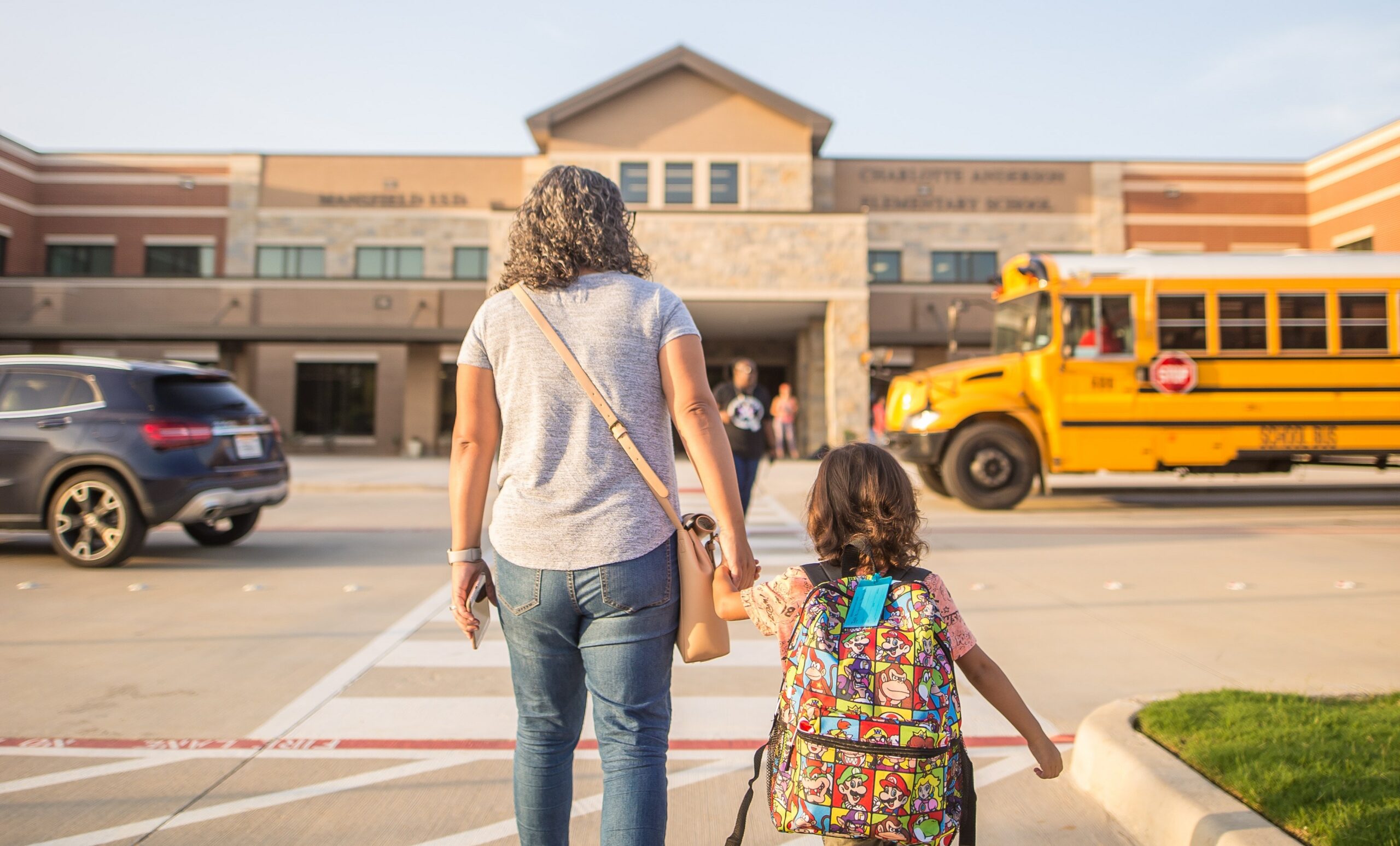 a student and parent are walking into a school building. A school bus is parked off to the right and a car is driving away on the left.