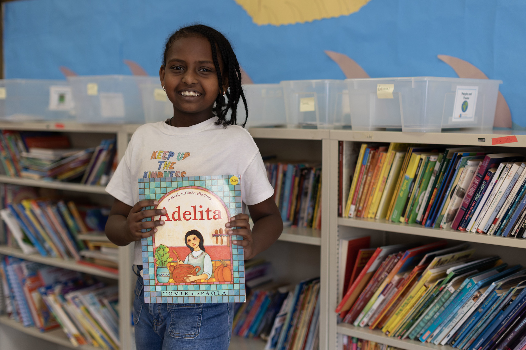 student holding a book, smiling in front of a bookshelf stacked with more books libraries