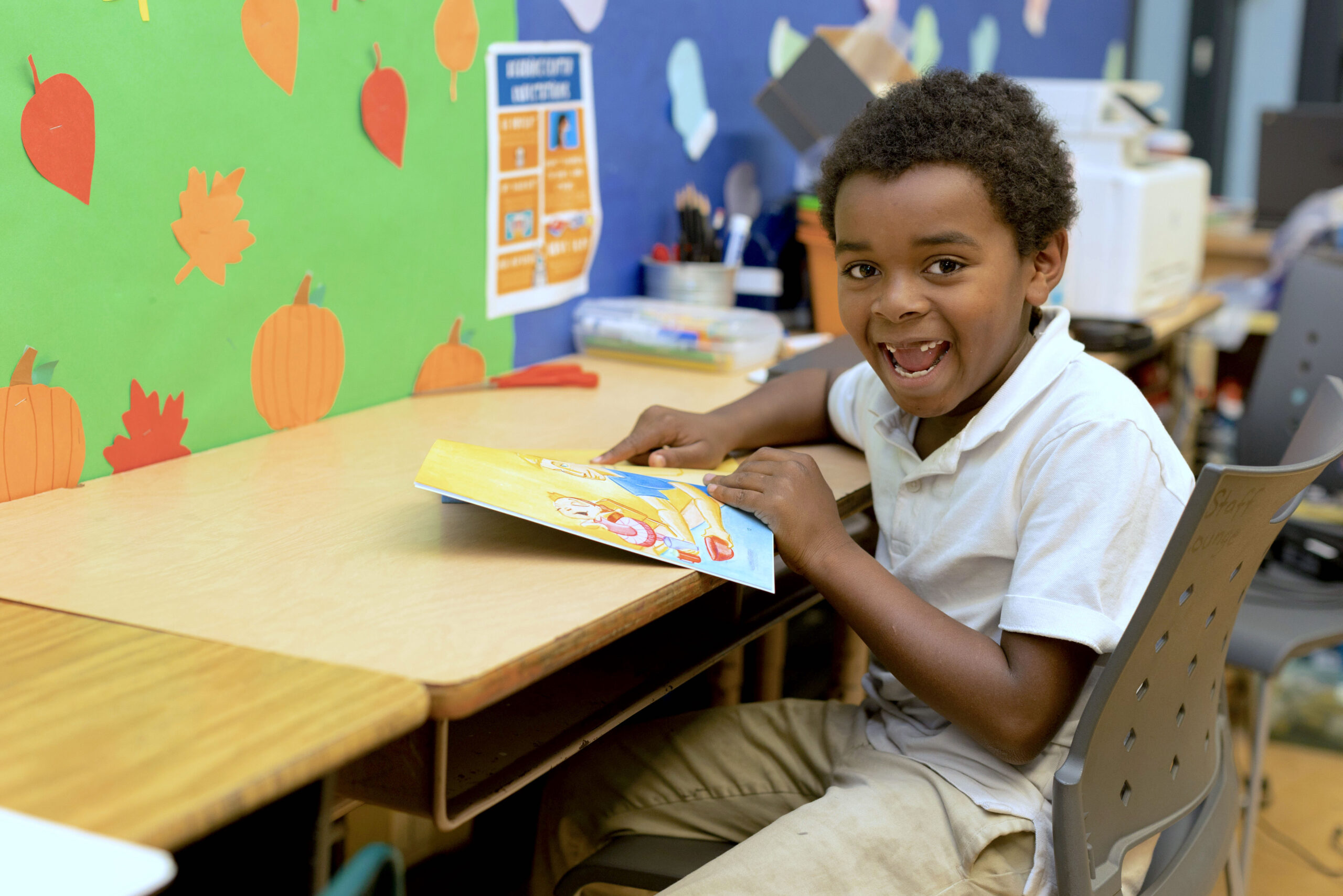 male student sitting at a desk, pointing to a page on a book, smiling at camera