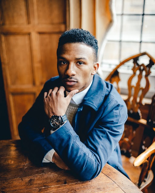 brandon harris poses in a suit with one hand on his chin, he's seated in a well-lit room on a wooden chair, americorps service term