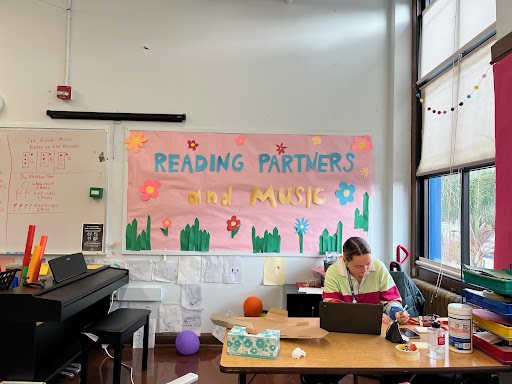 an americorps member serving at a desk, behind her is a sign that reads "Reading Partners and music," americorps service term