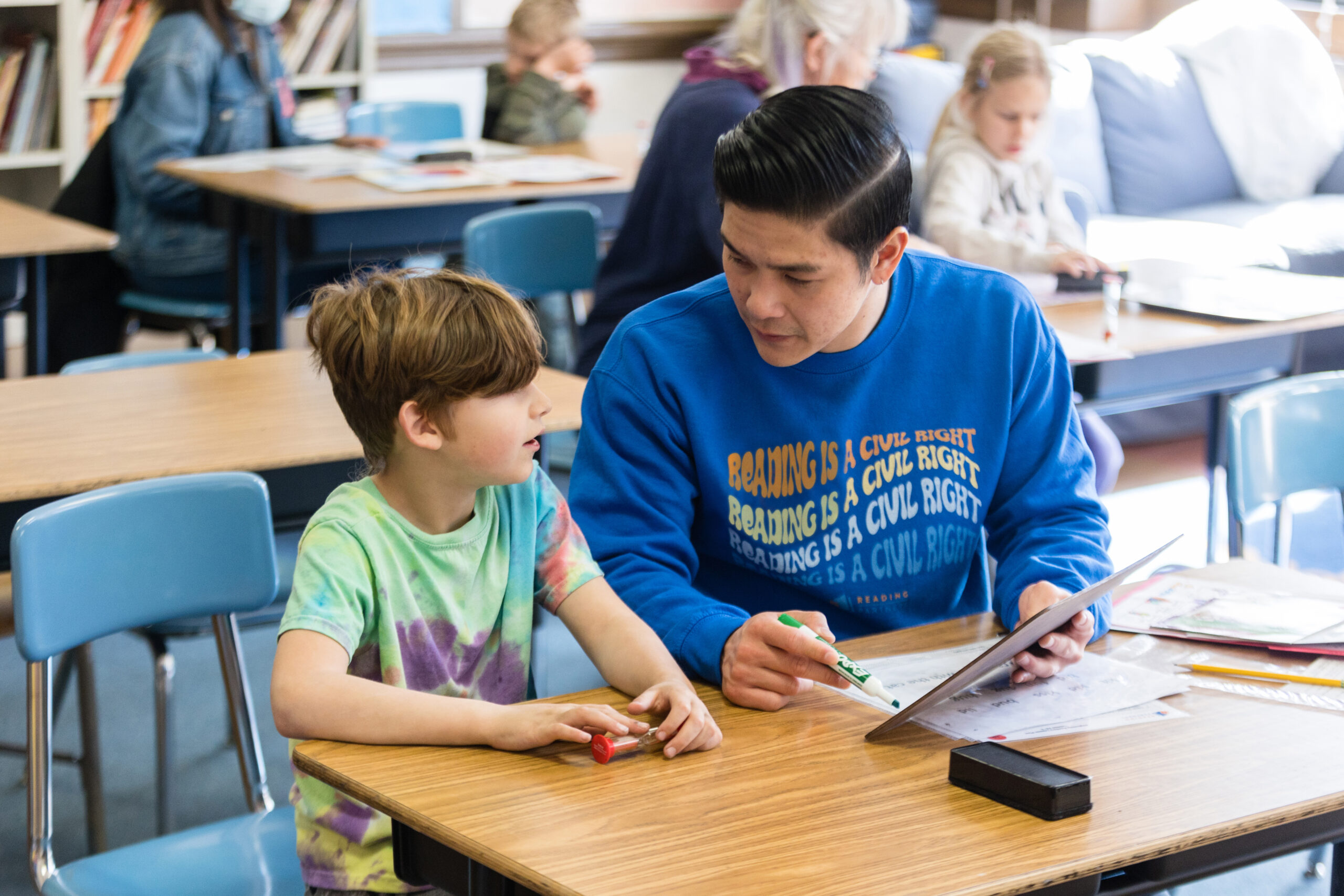 tutor and student talking at a desk in a classroom, the tutor wears a sweatshirt that says "Reading is a civil right"