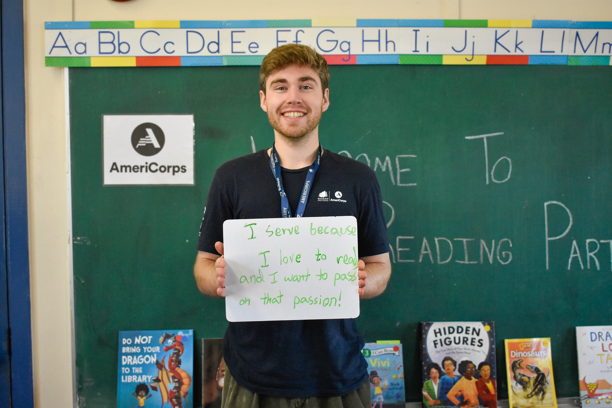 an americorps member holds a sign that reads "I serve because I love to read and I want to pass on that passion," recruiting americorps members