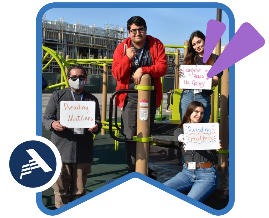 four americorps members on a playground, smiling at the camera and holding up whiteboards that read "Reading matters" and "Laughter keeps us going"