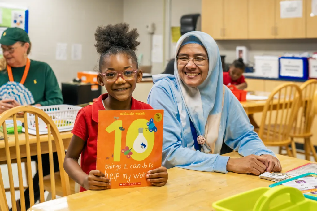 a tutor and student sitting at a table, the student holding up a book and smiling